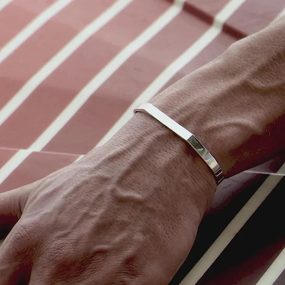 A close-up of a person’s wrist wearing a minimalist cuff bracelet. The hand rests on a surface with diagonal red and white stripes, and prominent veins are visible on the hand and wrist.