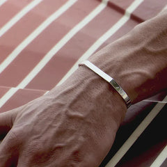 A close-up of a person’s wrist wearing a minimalist cuff bracelet. The hand rests on a surface with diagonal red and white stripes, and prominent veins are visible on the hand and wrist.