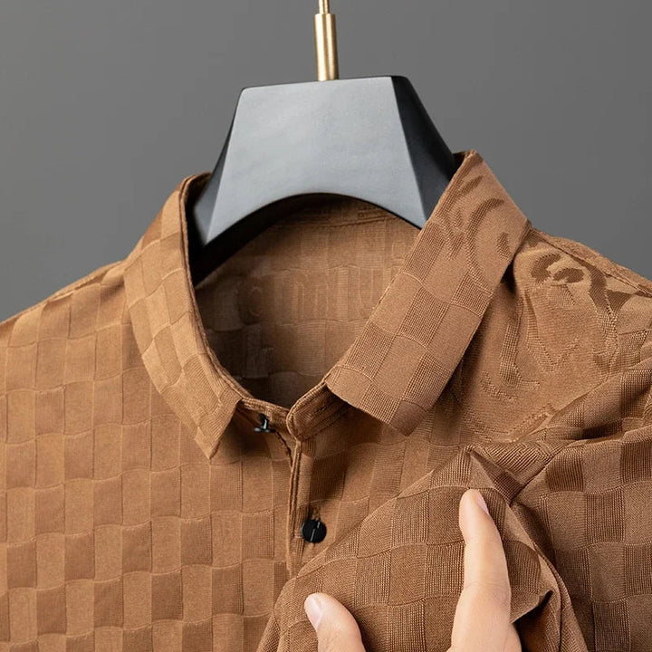 A brown checkered shirt in a tailored fit hangs on a hanger, with a hand lifting the fabric to reveal its silk cotton blend texture and checkerboard pattern. The shirt features a buttoned collar and a subtle shiny finish.