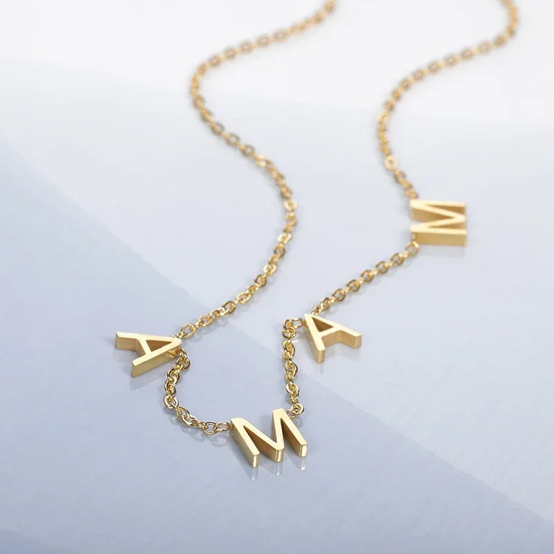 A gold stainless steel necklace with three dangling letter charms spelling MAMA, styled as minimalist jewelry, displayed on a light blue and white background.