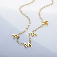A gold stainless steel necklace with three dangling letter charms spelling MAMA, styled as minimalist jewelry, displayed on a light blue and white background.