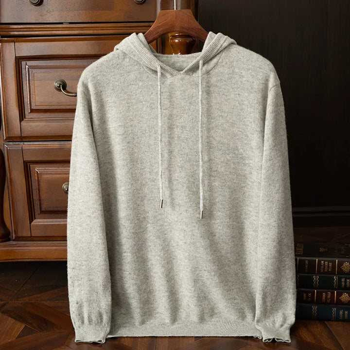 A light gray cashmere blend hooded sweater with drawstrings hangs on a wooden hanger, placed in front of a wooden dresser. Some books are partially visible on the right side of the image.