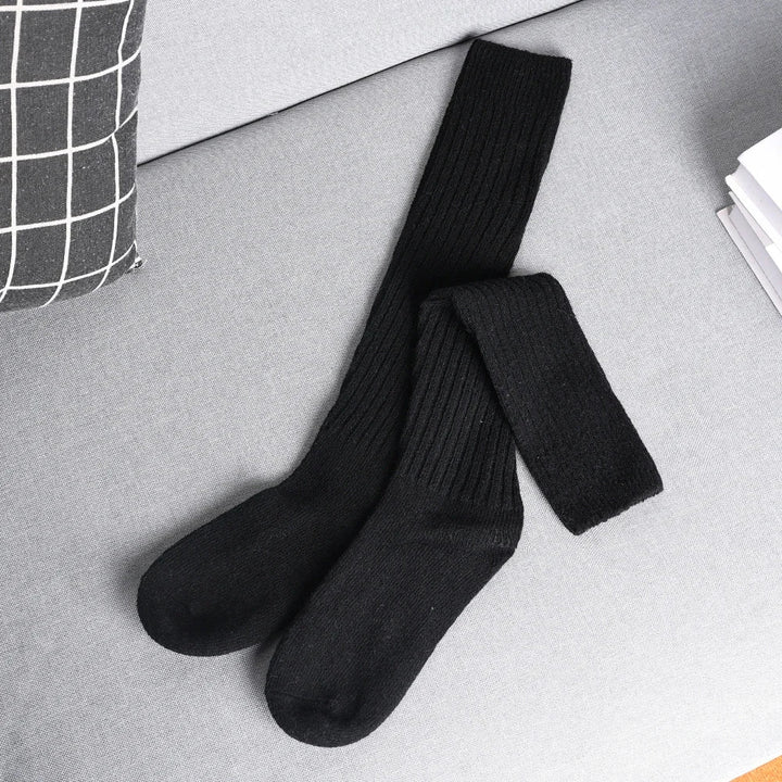 A pair of long, black, ribbed socks made from a Cashmere cotton blend are laid out on a light grey sofa next to a black and white checkered pillow and a stack of white papers.