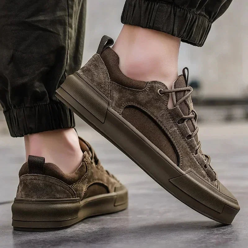 A person in dark green jogger pants and stylish brown suede canvas shoes with cushioned insoles stands on a smooth gray surface. The sneakers feature textured suede details, lace-up fronts, and thick, durable rubber outsoles for added comfort.