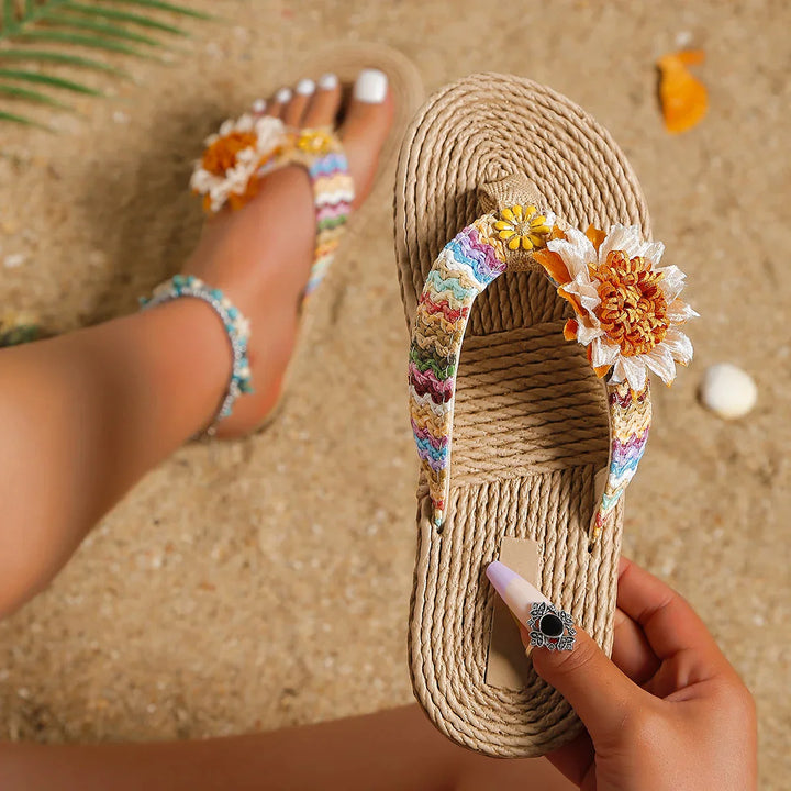 A person holds a woven sandal with colorful zigzag straps and a large fabric flower, while wearing a matching sandal with an anti-slip sole on the other foot—showcasing a fun women's fashion accessory on sandy ground.