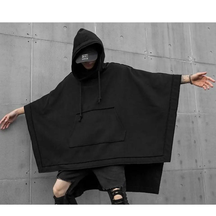 A person wearing a black techwear hoodie poncho and black cap poses with arms outstretched against a gray concrete wall. The streetwear outfit features a large front pocket and distressed black shorts.