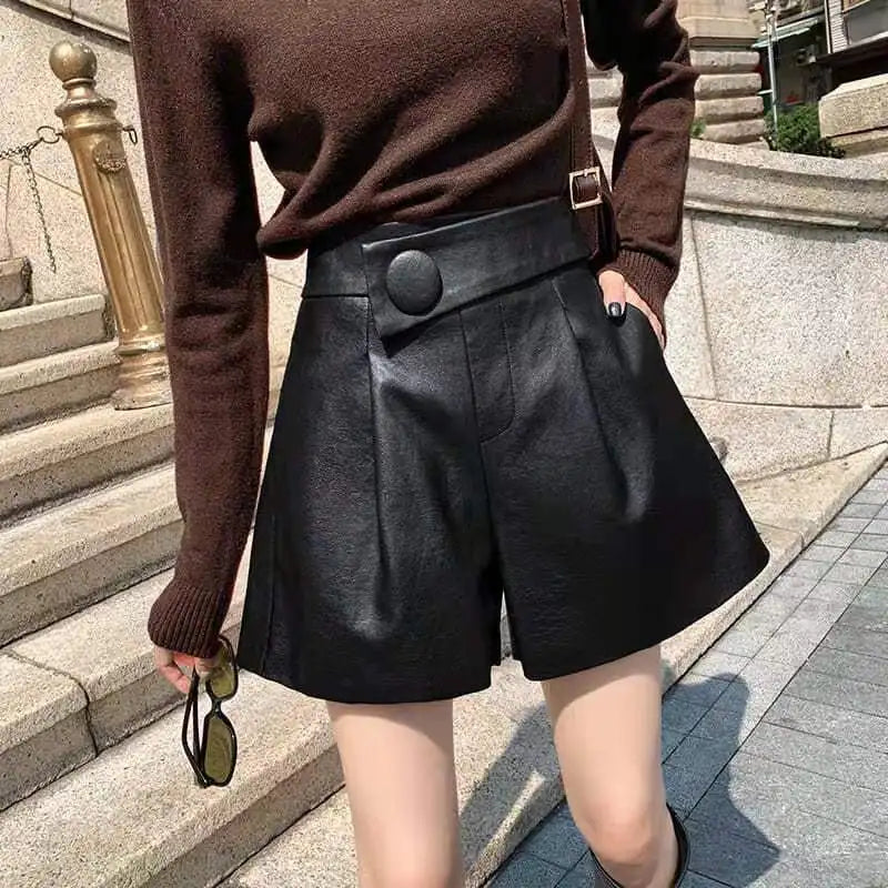 A person stands outdoors on stone steps wearing a brown long-sleeve top and high waist black calfskin leather shorts with button detail, holding sunglasses in one hand.