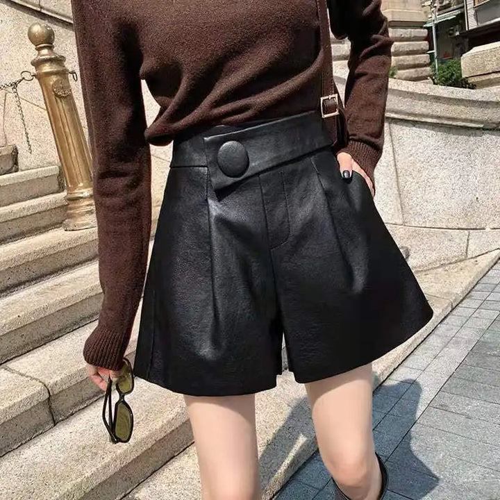 A person stands outdoors on stone steps wearing a brown long-sleeve top and high waist black calfskin leather shorts with button detail, holding sunglasses in one hand.