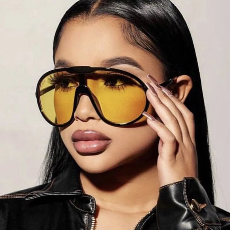 A woman with long straight black hair and glossy lips wears oversized sunglasses with UV400 protection and a black jacket, holding the glasses with one hand while looking to the side.