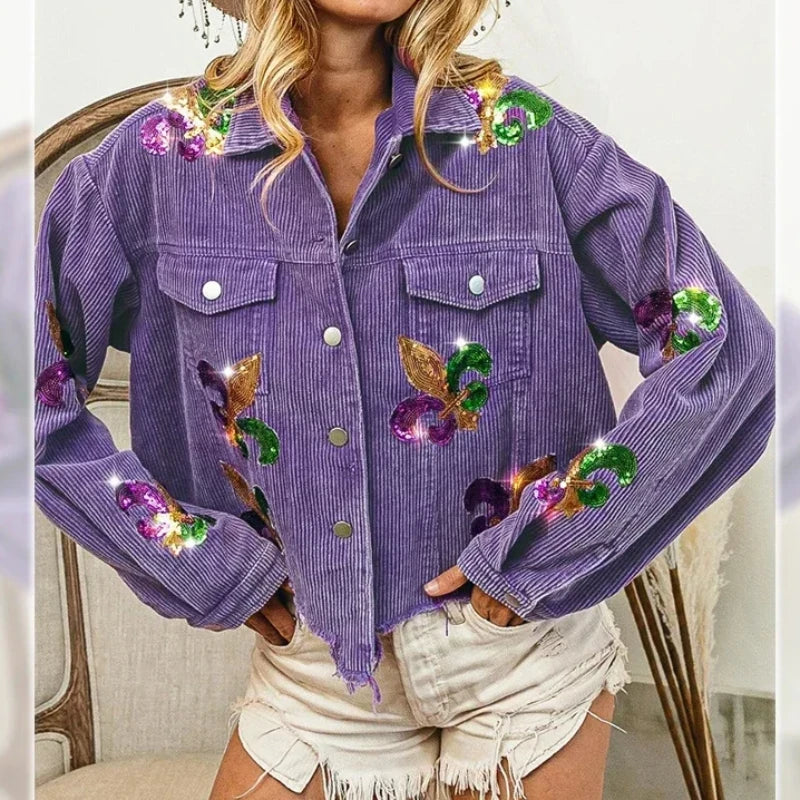 A woman wears a purple corduroy jacket with sequin floral patches and silver buttons, paired with light-colored, frayed denim shorts. Her hands rest on her hips.