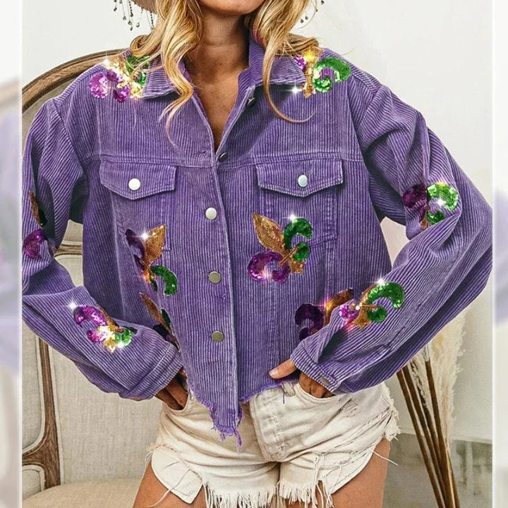 A woman wears a purple corduroy jacket with sequin floral patches and silver buttons, paired with light-colored, frayed denim shorts. Her hands rest on her hips.