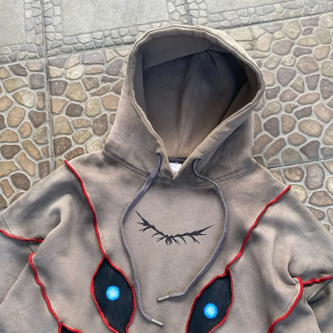 A gray hooded sweatshirt with red and black stitched designs resembling large, blue-highlighted eyes and dark, branch-like lines on the chest features eye pattern patchwork, laid out on a stone-patterned surface.