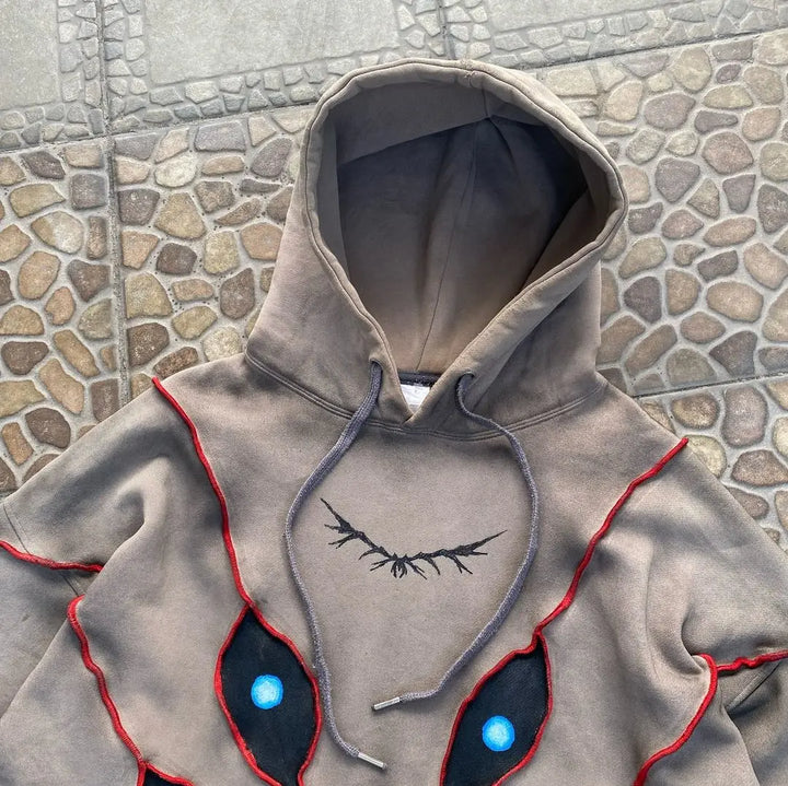 A gray hooded sweatshirt with red and black stitched designs resembling large, blue-highlighted eyes and dark, branch-like lines on the chest features eye pattern patchwork, laid out on a stone-patterned surface.