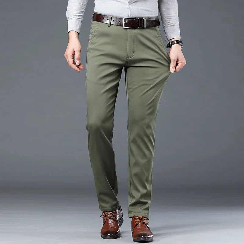 Person wearing olive green pants with a tailored fit, a light gray tucked-in shirt, dark brown belt, and brown dress shoes stands against a plain gray background. Their versatile style is perfect for any occasion, hands relaxed by their sides.