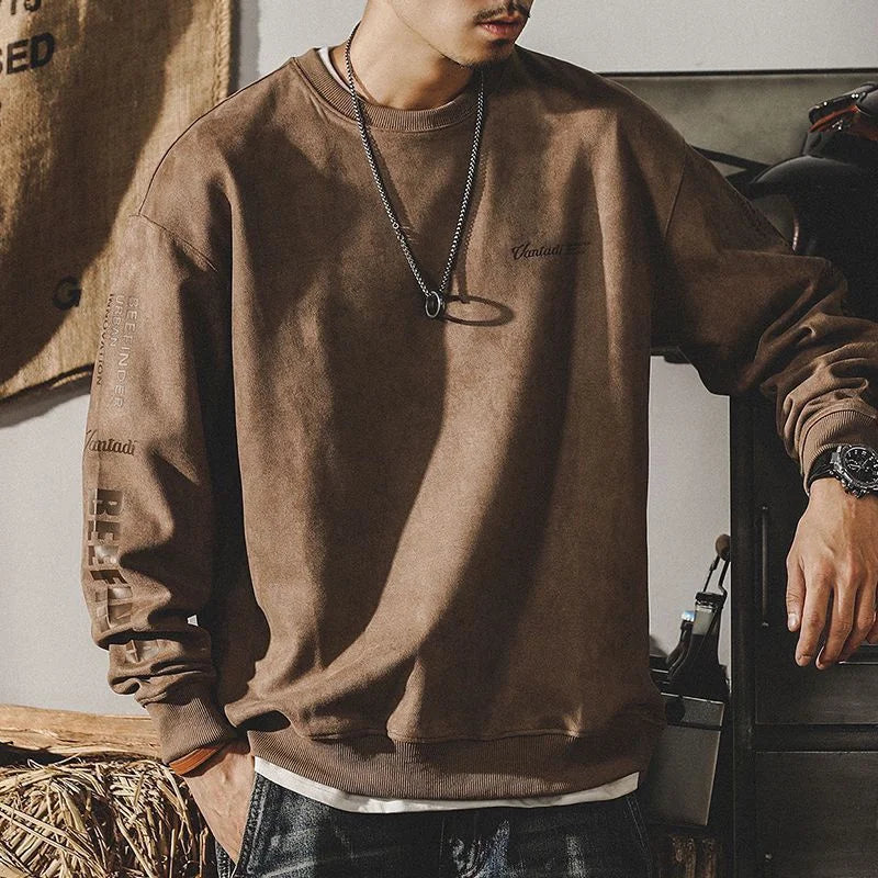 A person wearing a relaxed fit crewneck brown sweatshirt with printed text on the sleeves and chest, paired with a necklace, watch, and jeans, stands indoors with their hand resting on a surface. Their face is partially out of frame.