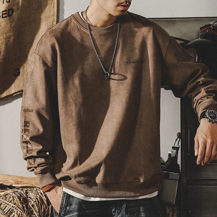 A person wearing a relaxed fit crewneck brown sweatshirt with printed text on the sleeves and chest, paired with a necklace, watch, and jeans, stands indoors with their hand resting on a surface. Their face is partially out of frame.