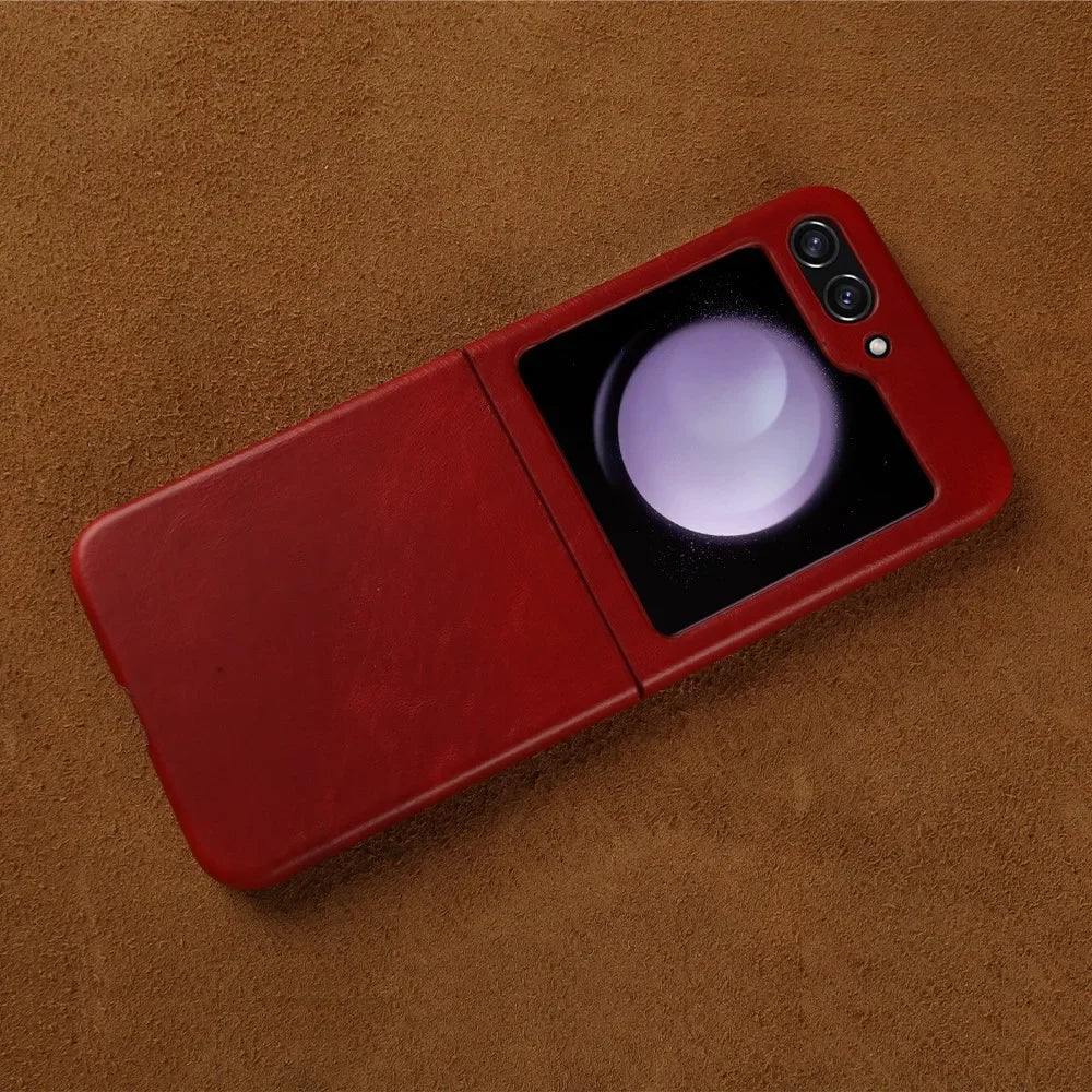 A red genuine leather flip phone case designed for Samsung Galaxy Z Flip rests on a brown suede surface. The phone is partially folded, its external screen aglow, and dual camera lenses visible—offering both style and shockproof protection.