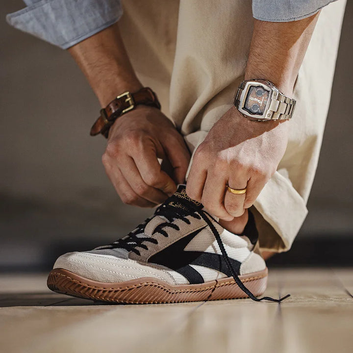 A person wearing light pants and a wristwatch is kneeling down, tying the laces of beige and black Olympism 1984 Pro Sneakers with a gum sole, channeling 80s aesthetics on a wooden floor.