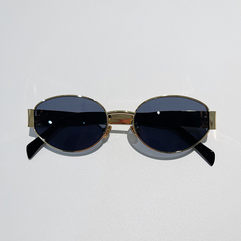 Oval-shaped, vintage-inspired sunglasses with gold metal frames and dark tinted lenses offer UV400 protection. Placed on a light gray surface, the arms of these stylish glasses are slightly angled outward.
