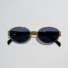 Oval-shaped, vintage-inspired sunglasses with gold metal frames and dark tinted lenses offer UV400 protection. Placed on a light gray surface, the arms of these stylish glasses are slightly angled outward.