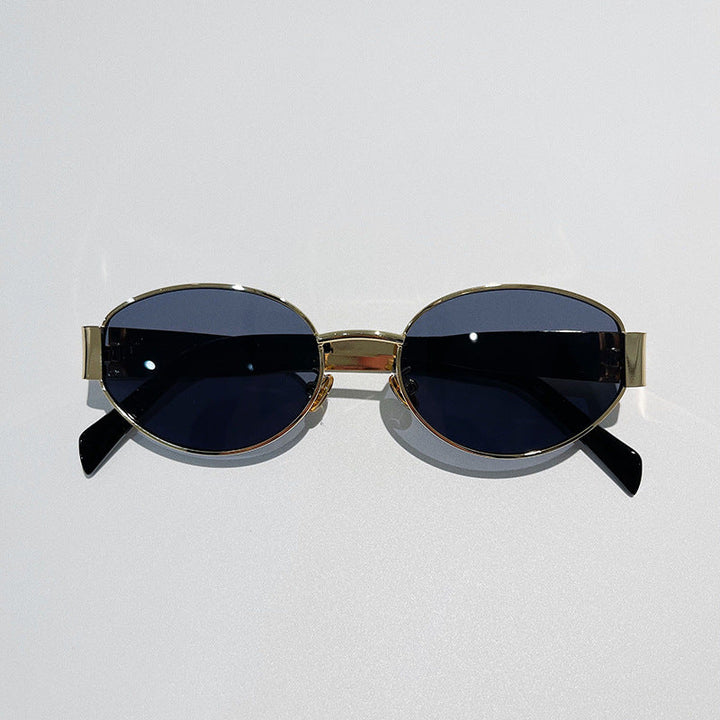 Oval-shaped, vintage-inspired sunglasses with gold metal frames and dark tinted lenses offer UV400 protection. Placed on a light gray surface, the arms of these stylish glasses are slightly angled outward.