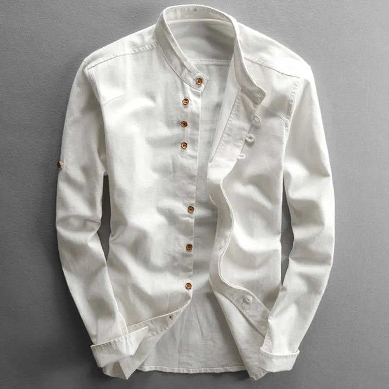 A white, long-sleeved Japanese inspired overshirt with a stand mandarin collar and wooden buttons arranged asymmetrically down the front. Displayed against a gray background, this 100% cotton shirt has a casual, slightly wrinkled look.
