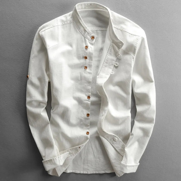 A white, long-sleeved Japanese inspired overshirt with a stand mandarin collar and wooden buttons arranged asymmetrically down the front. Displayed against a gray background, this 100% cotton shirt has a casual, slightly wrinkled look.
