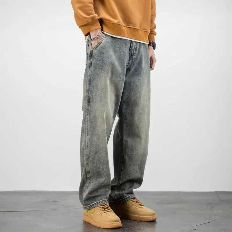 A person wearing a brown sweatshirt, classic denim straight leg jeans, and tan sneakers stands with hands in pockets against a plain light-colored background.