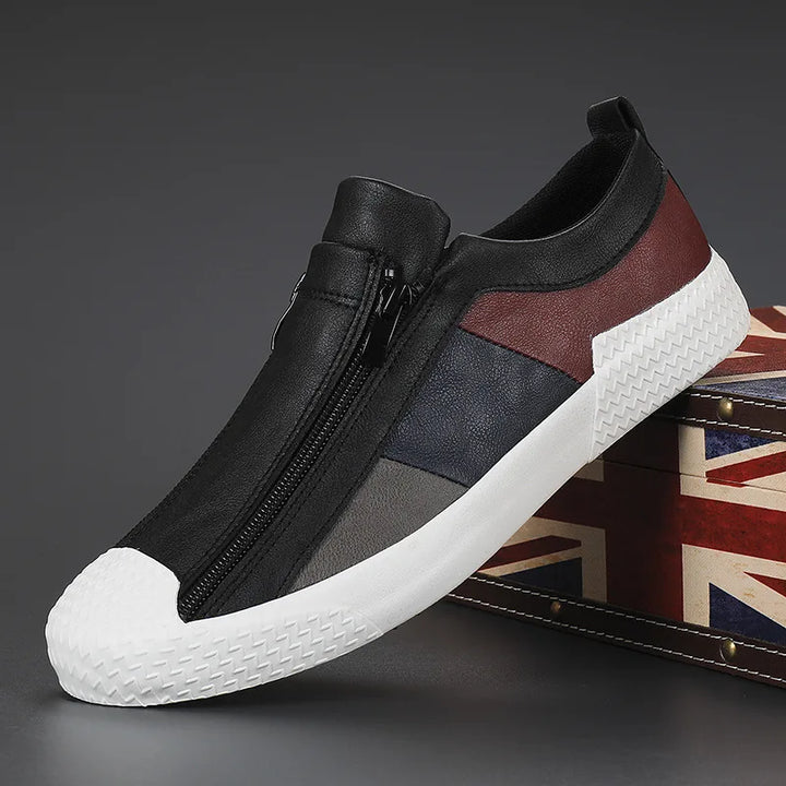 A stylish sneaker with a white textured, cushioned sole, black zipper closure, and panels of black, gray, navy, and burgundy. The shoe is displayed leaning against a box with a British flag design on a dark background.