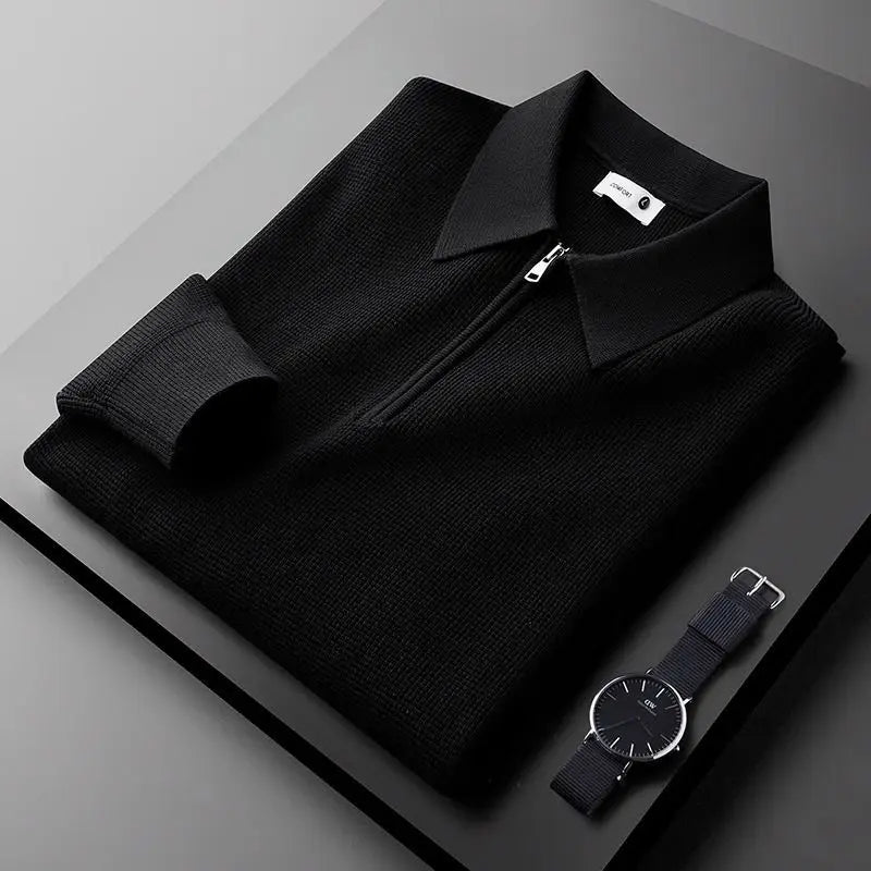 A neatly folded black zip-up sweater made from stretchy fabric with a collar is placed on a dark surface next to a minimalist black wristwatch with a fabric strap.