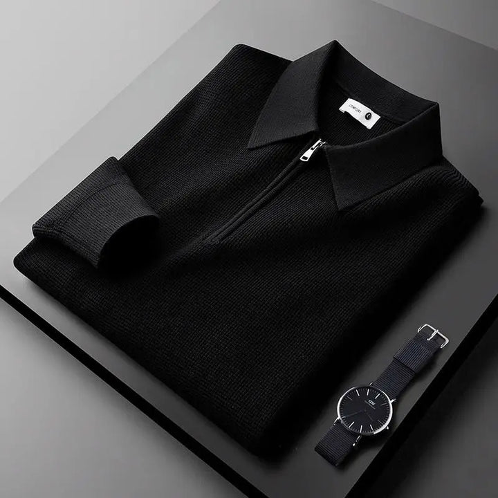 A neatly folded black zip-up sweater made from stretchy fabric with a collar is placed on a dark surface next to a minimalist black wristwatch with a fabric strap.