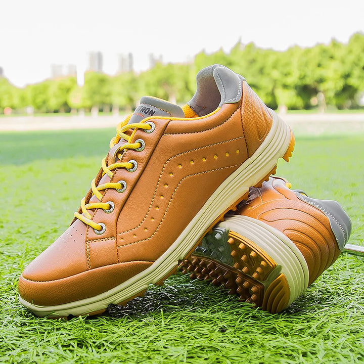 A pair of tan and yellow athletic sneakers with white soles, displayed on green grass outdoors with trees and buildings blurred in the background. Showcasing advanced cleat technology, this golf footwear combines style and performance.