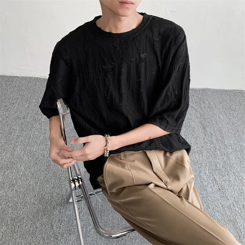 A person sits on a clear chair, wearing a textured black oversized tee and beige pleated pants, arms resting casually with a bracelet on one wrist. The background is plain with grey carpet and a white wall, evoking laid-back streetwear vibes.