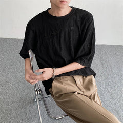 A person sits on a clear chair, wearing a textured black oversized tee and beige pleated pants, arms resting casually with a bracelet on one wrist. The background is plain with grey carpet and a white wall, evoking laid-back streetwear vibes.
