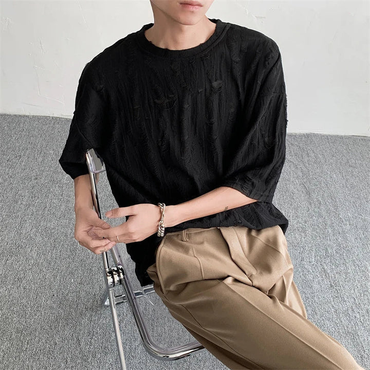 A person sits on a clear chair, wearing a textured black oversized tee and beige pleated pants, arms resting casually with a bracelet on one wrist. The background is plain with grey carpet and a white wall, evoking laid-back streetwear vibes.