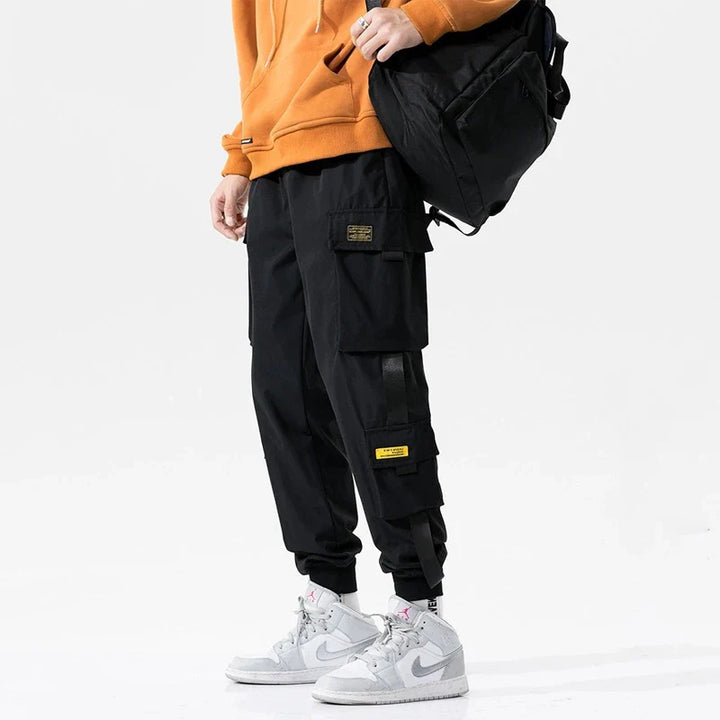 A person wearing a bright orange hoodie, black cargo pants with ribbed ankle cuffs and an elastic waist, yellow patches, white Nike sneakers, and carrying a black backpack, stands against a plain white background.