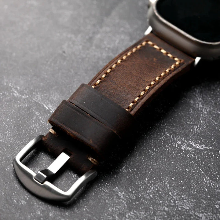 Close-up of a handcrafted brown Italian cowhide leather watch strap with beige saddle stitched detailing and a silver buckle, attached to a metal watch case, resting on a textured dark surface.