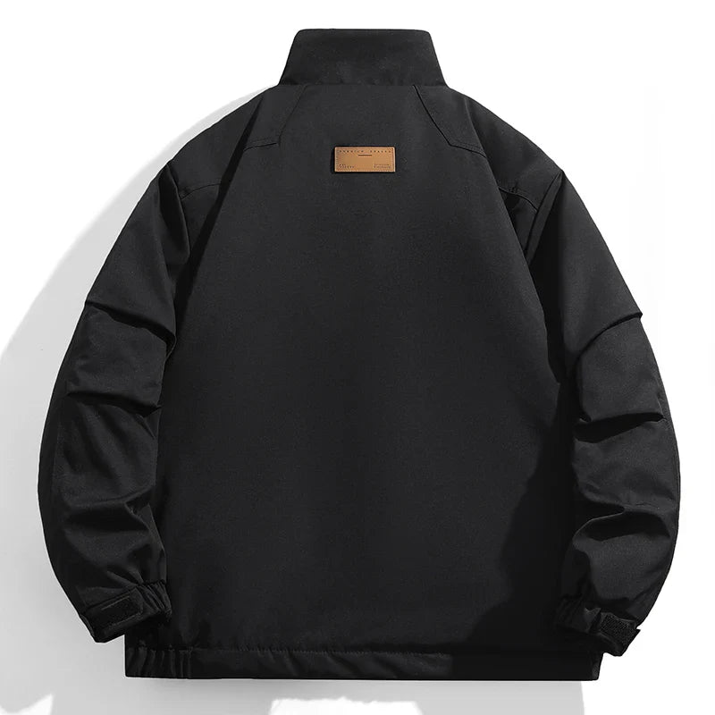 Black windproof jacket viewed from the back, featuring a high collar, adjustable cuffs, and a small rectangular brown patch near the upper center of the back. The background is white.