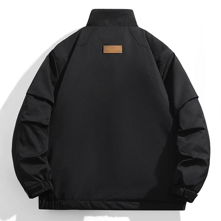 Black windproof jacket viewed from the back, featuring a high collar, adjustable cuffs, and a small rectangular brown patch near the upper center of the back. The background is white.