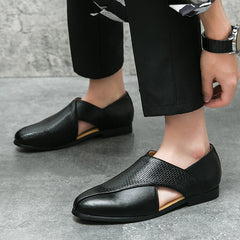 A person wears black genuine leather loafers with textured and smooth panels and side cutouts. Paired with black pants and a watch, they stand on a gray floor against a concrete wall.