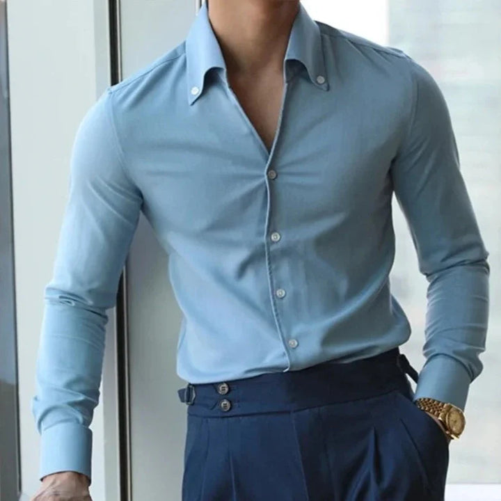 A person wearing a light blue wrinkle resistant shirt and dark blue high-waisted trousers stands near a window, one hand in their pocket and a gold watch on their wrist. Their face is not visible.
