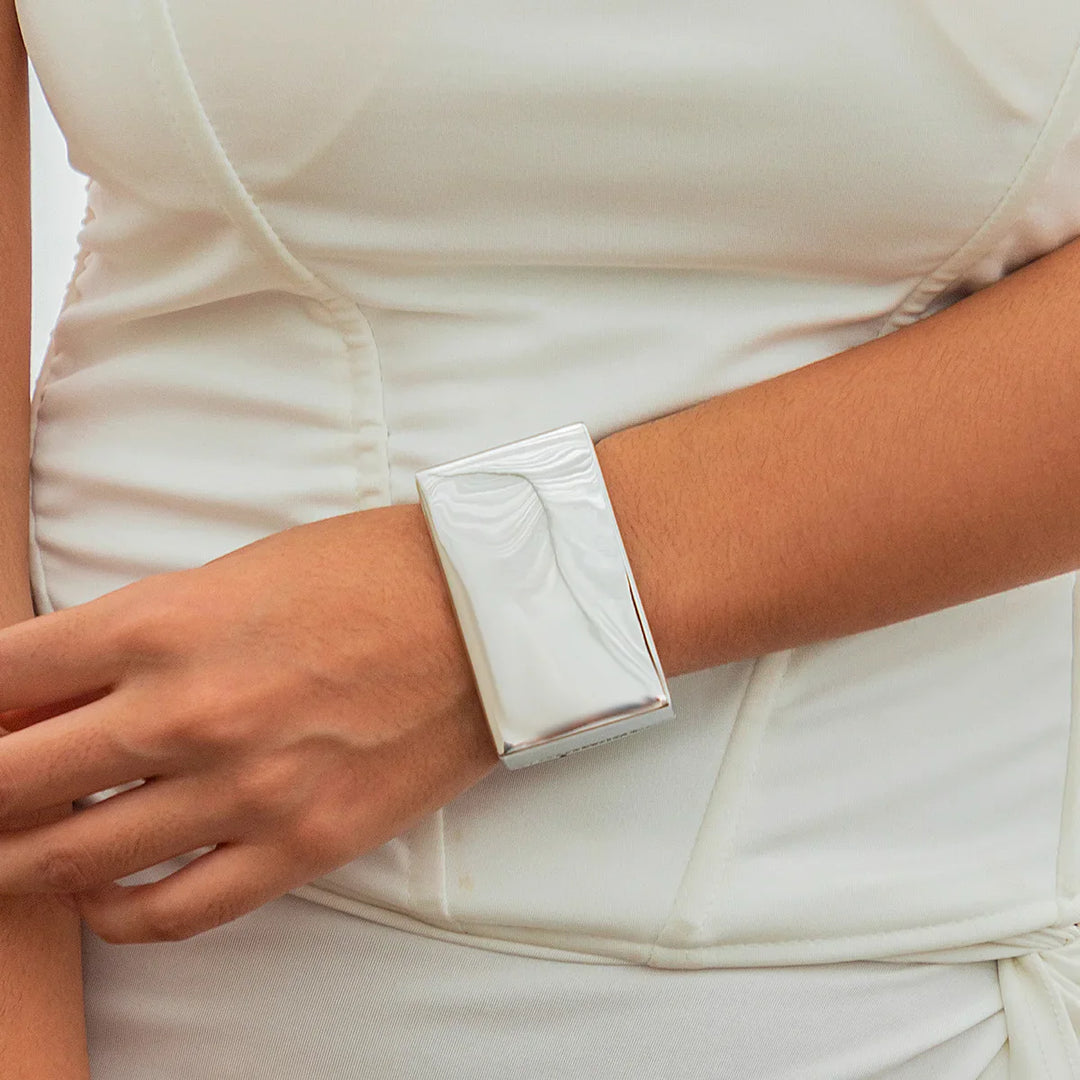 A person wearing a white, fitted dress accessorizes with a large, shiny, rectangular silver cuff bracelet featuring a bold geometric design on their wrist. Their hand rests gently near their waist.