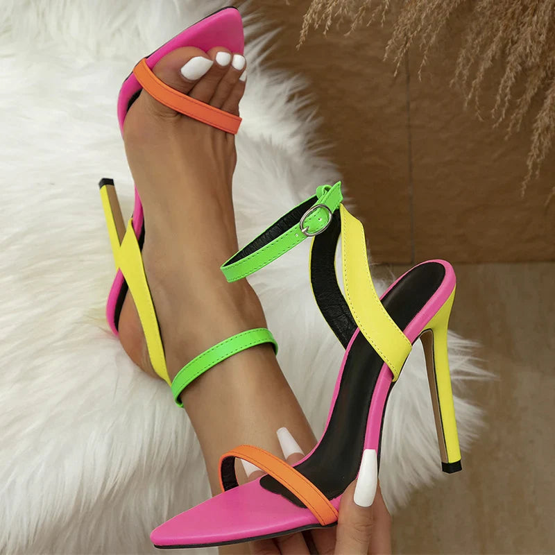 A person wearing fashion-forward, high-heeled sandals with neon pink, orange, yellow, and green straps. The pointed-toe shoes feature stiletto heels and an adjustable ankle strap. White-painted nails rest on a plush white furry rug.