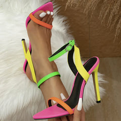 A person wearing fashion-forward, high-heeled sandals with neon pink, orange, yellow, and green straps. The pointed-toe shoes feature stiletto heels and an adjustable ankle strap. White-painted nails rest on a plush white furry rug.