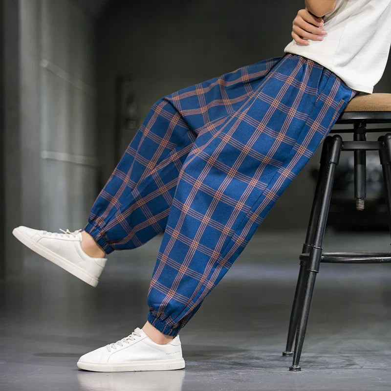 A person sits sideways on a stool in a minimal indoor setting with grey walls and floors, wearing loose blue plaid cotton joggers with elastic cuffs and white sneakers—a modern nod to Tokyo streetwear.