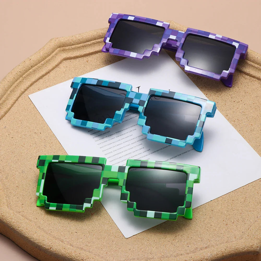 Three pairs of pixelated sunglasses in purple, blue, and green with chunky, block-like frames channel a retro-gaming aesthetic as they rest on a beige tray with white paper underneath.