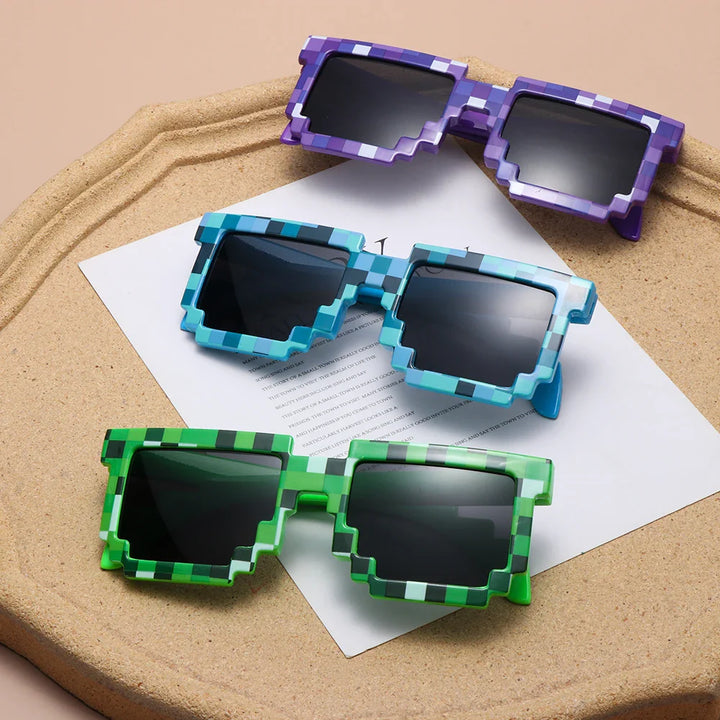 Three pairs of pixelated sunglasses in purple, blue, and green with chunky, block-like frames channel a retro-gaming aesthetic as they rest on a beige tray with white paper underneath.
