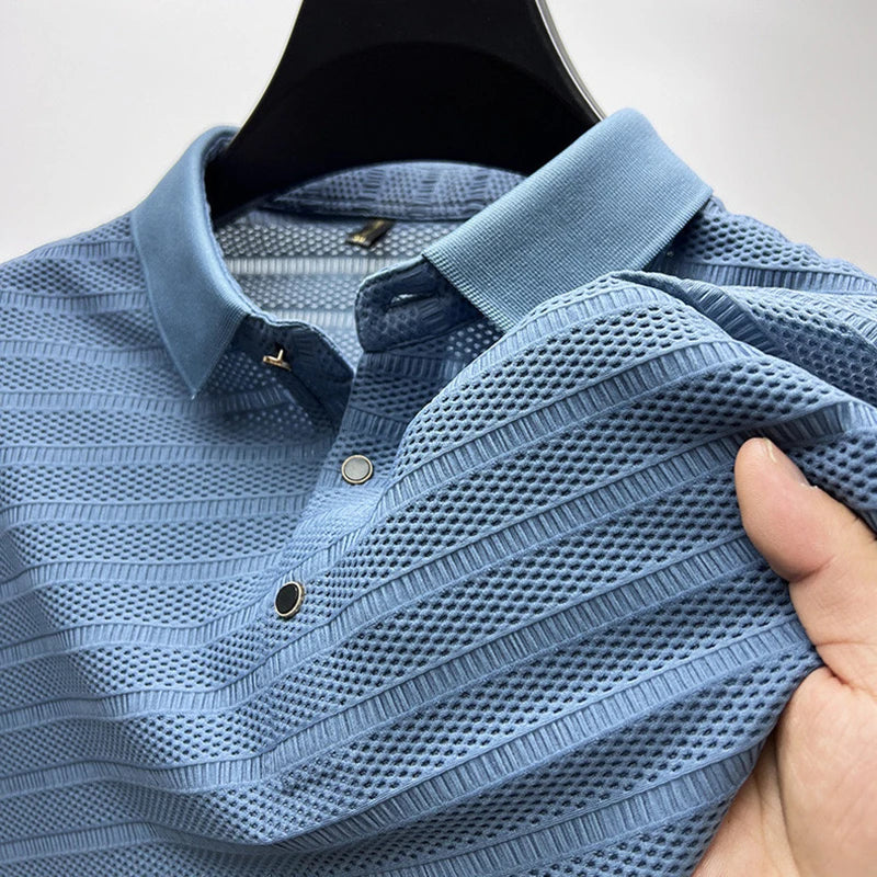 A close-up of a light blue, textured button-up shirt on a hanger. A hand lifts the shirt to reveal its silk fabric, highlighting mesh-like breathability and detailed stitching—perfect for a classic polo look.