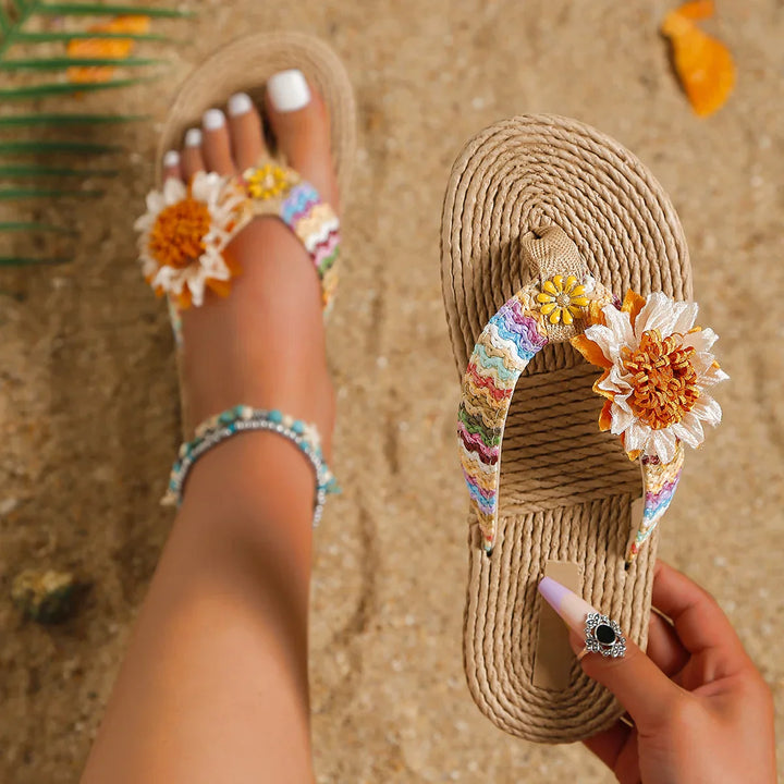 A person wearing woven sunflower flip flops with colorful straps and decorative fabric flowers, sitting on sandy ground. One sandal—a trendy women's fashion accessory—is held by a hand with pastel nails and a ring, revealing a beaded anklet.