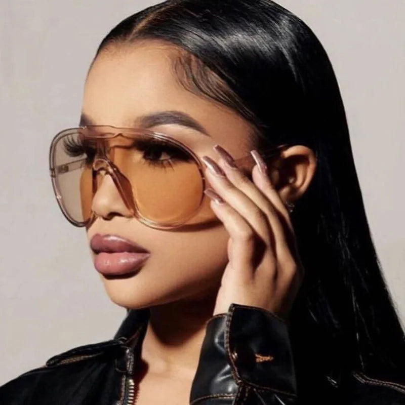 A woman with long, straight black hair wears oversized, amber-tinted sunglasses with UV400 protection and a black leather jacket. She holds the side of her glasses and looks confidently to the left.